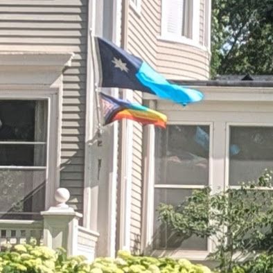 Minnesota state flag flies,with a Progress Pride flag, from a flagpole in front of a cream colored house in Saint Paul, MN