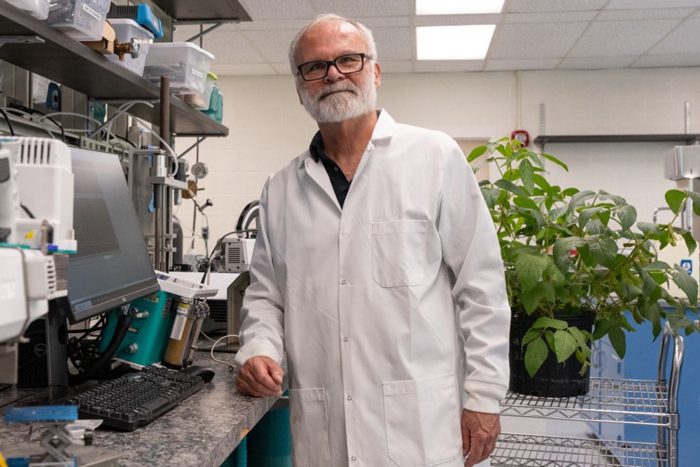 Tom Sharkey poses in a lab coat in front of a plant and computer in his lab.