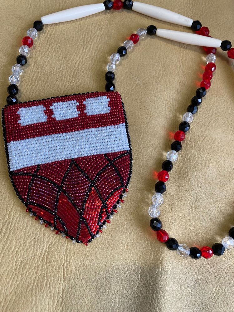 shows a beaded medallion laying on a piece of deer hide. the necklace of the medallion consists of matte/shiny black, red, and clear fire polish beads and bone beads. the medallion is of the harvard law school shield. with maroon beads of different finishes on the bottom of the shield, a stripe of white translucent czech seed beads in the middle, and maroon beads surrounding white rectangles at the top of the shield. it is edged in majority black beads, with an alternating red and white pattern at the bottom of the shield. 