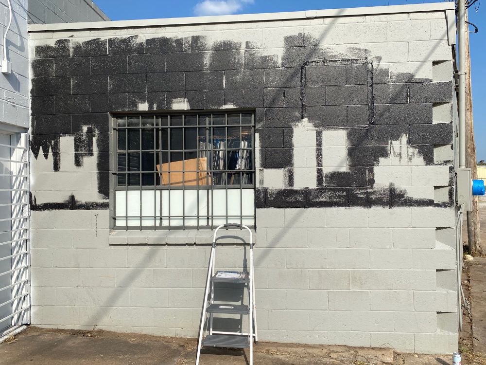gray block wall with a caged window in the semi-center of it. the upper half of the wall is painted black, with various vague outlines in the black. there’s a step ladder in front of the window. 