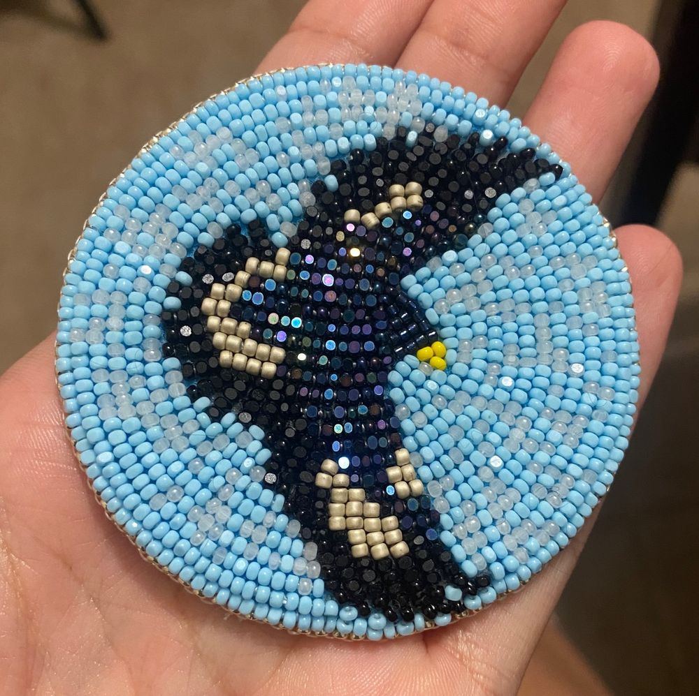 a circular piece of beadwork.
in the center of the beadwork is a beaded eagle, with black feathers. it’s beak is yellow and body iridescent black beads. the background is sky blue and cloudy translucent white beads. 