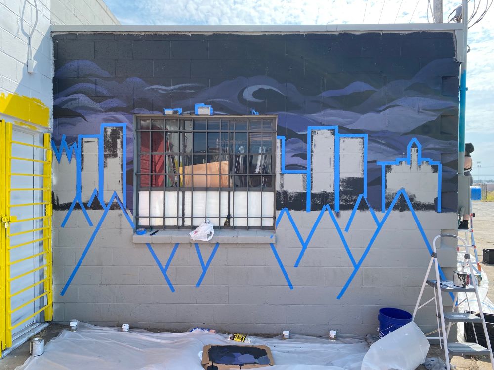 grey block wall. half painted black with swirling shades of blue and grey, depicting clouds. blue painters tape outlines shapes on both the black and grey half of the wall. 