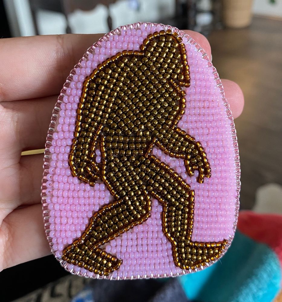 shows a close up of an arch shaped beaded piece. it’s background is mostly bubblegum pink opaque beads with a shiny metallic brown silhouette of bigfoot in the center. it is edged in translucent pink silver lined beads. 