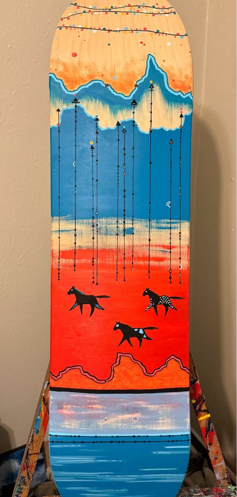 shows a painted skate deck propped on an easel vertically. the bottom is painted in varying shades of blue. above the truck holes is a thin black line. above that is cadmium red up to the middle of the board. there are three ledger style horses running on the red in a triangle formation. the top half of the board is the same color blue as the majority of the bottom, but depicting the silhouette of Badlands rock formations. behind the painted silhouette is the boards natural wood grain with painted circles depicting stars strung in the sky.