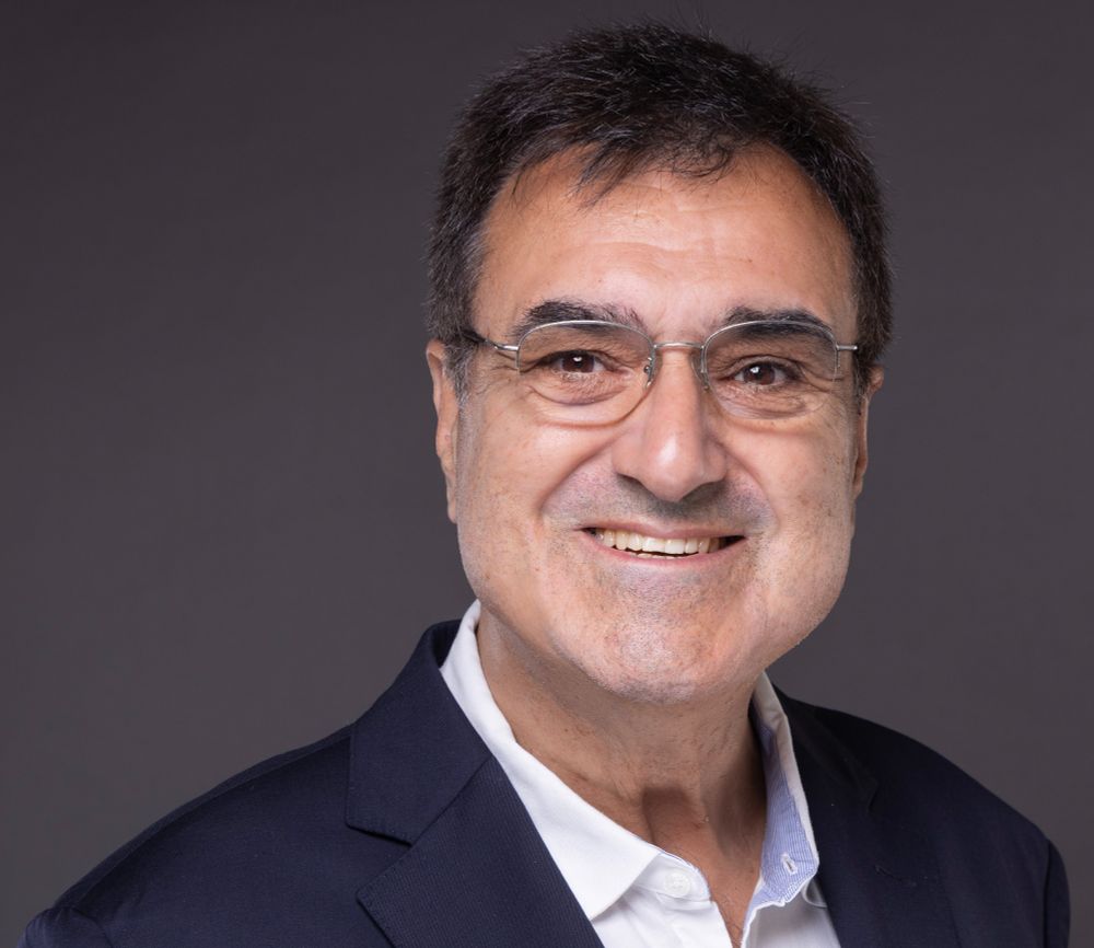 Portrait of Joaquim Jorge, smiling warmly in a dark blazer and white shirt, facing the camera against a neutral gray background. He is wearing glasses and exudes a friendly, professional demeanor.