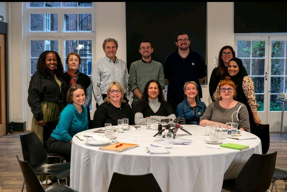 A group of 12 people facing the camera in a posed way. Smiling and pushed together - some are standing whilst others are sitting at a table. 