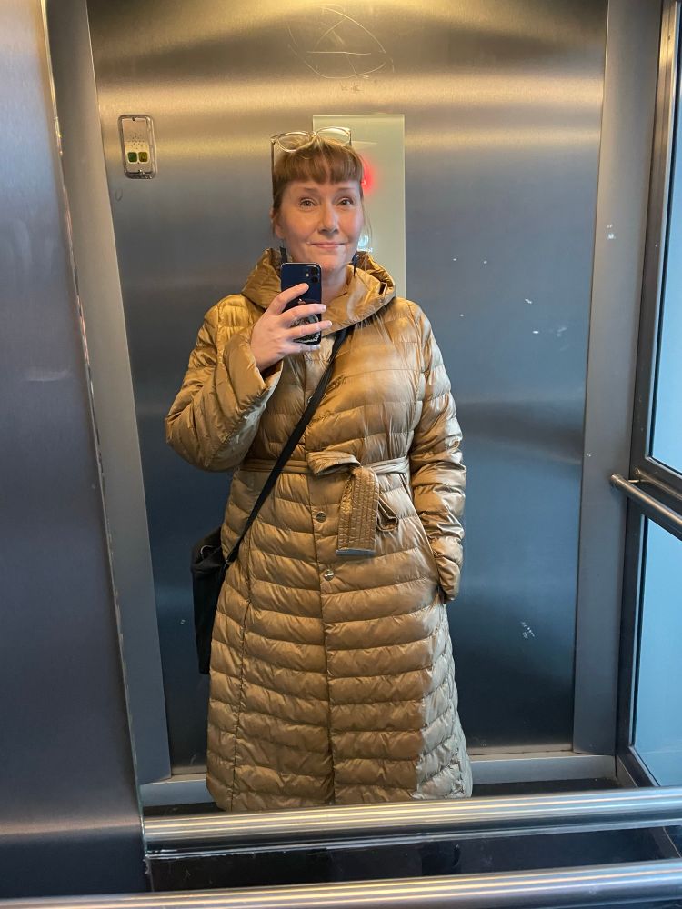 Hissiselfie keski-ikäisestä naisesta, joka on verhoutunut kullankeltaiseen kevytuntsikkaan. Hän hymyilee hieman. 