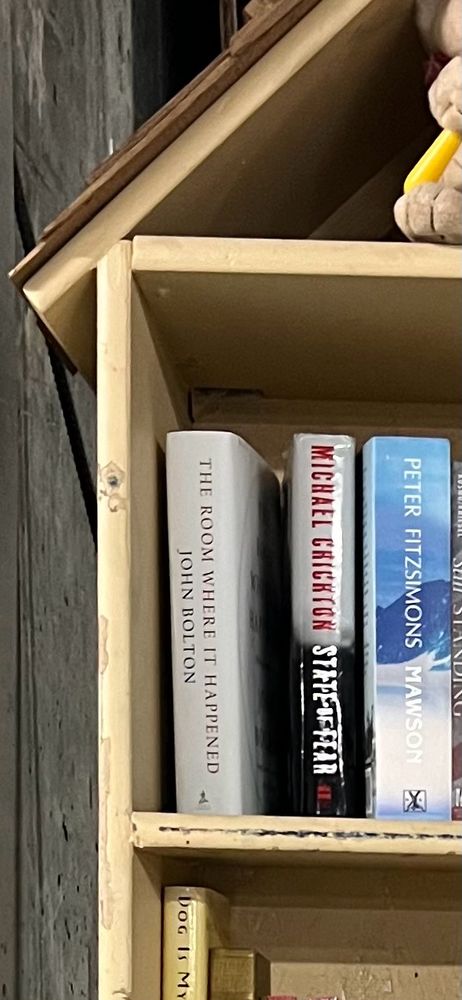An even closer view of a lending library in a wooden bookshelf. The bookshelf sits in the concrete-walled garage of a building in Big Sky, Montana. This photo centers the upper left corner of the top shelf, where three books are visible: The Room Where it Happened by John Bolton, State of Fear by Michael Crichton, and Mawson by Peter Fitzsimons.