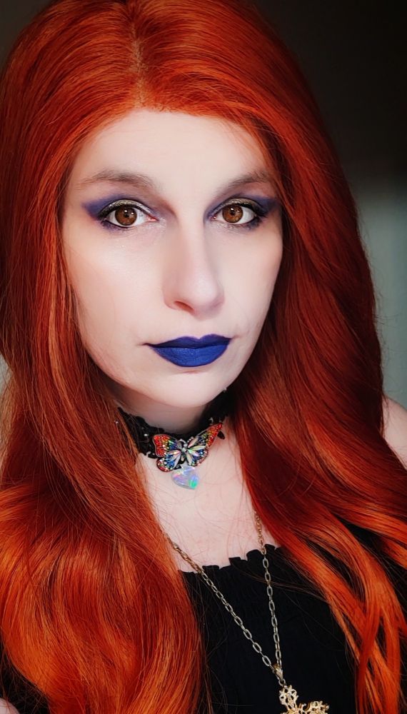 Selfie of a fem presenting trans person, with long, orange hair, wearing vibrant blue lipstick and eyeshadow. They also wear a black lace choker, with a rainbow butterfly piece, and a holographic heart charm, as well as an off the shoulder, black dress. 