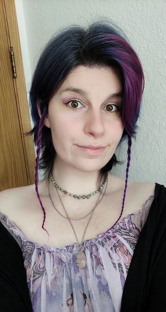 A selfie of a fem presenting, androgynous person with short, dark blue hair with light violet bangs and braided rat tails. They're wearing an off the shoulder lavender dress, and black cardigan, along with multiple handmade necklaces. One a plain chain, one a rose quartz pendant, the last a choker made of small labradorite stones. 