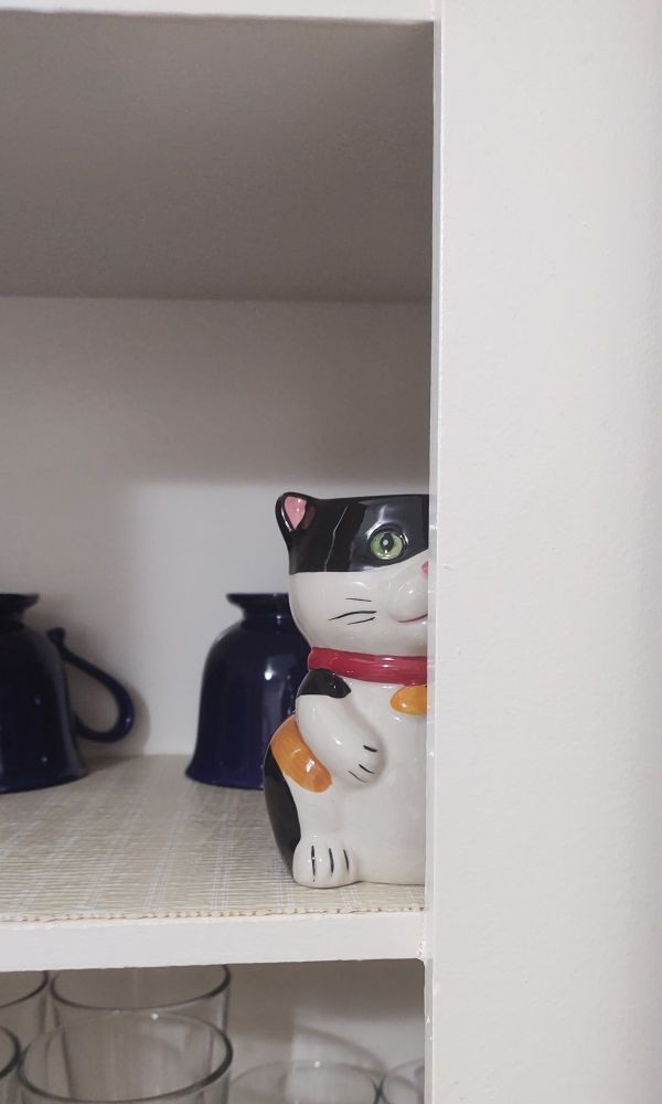 A mug in the appearance of a calico cat, wearing a red collar with a goldfish charm, peeking out behind the edge of a cabinet with several mugs behind it, the calico mug is, notably, the only one right side up. the mug appears to be spying on the rest of the kitchen. It may have a playful appearance when viewed in whole, but only part of its face is visible, giving it a slightly unsettling appearance. 