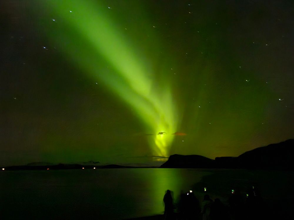 a green streak of aurora across the night sky, a lake in the foreground, some tourists with phones visible closer still