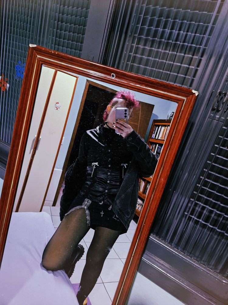 foto no espelho (apoiado em uma janela) mostrando uma pessoa branca com moicano roxo usando uma camisa de cetim com apliques de laço branco e duas espadinhas. usa também uma minissaia preta com uma fenda e apliques góticos 