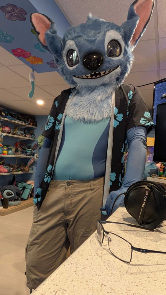 me leaning against my desk (that my phone taking the photo is sitting on), iʻm wearing a stitch fursuit head, bodysuit, cargo shorts, and black with blue flower patterned button up -- it is unbuttoned