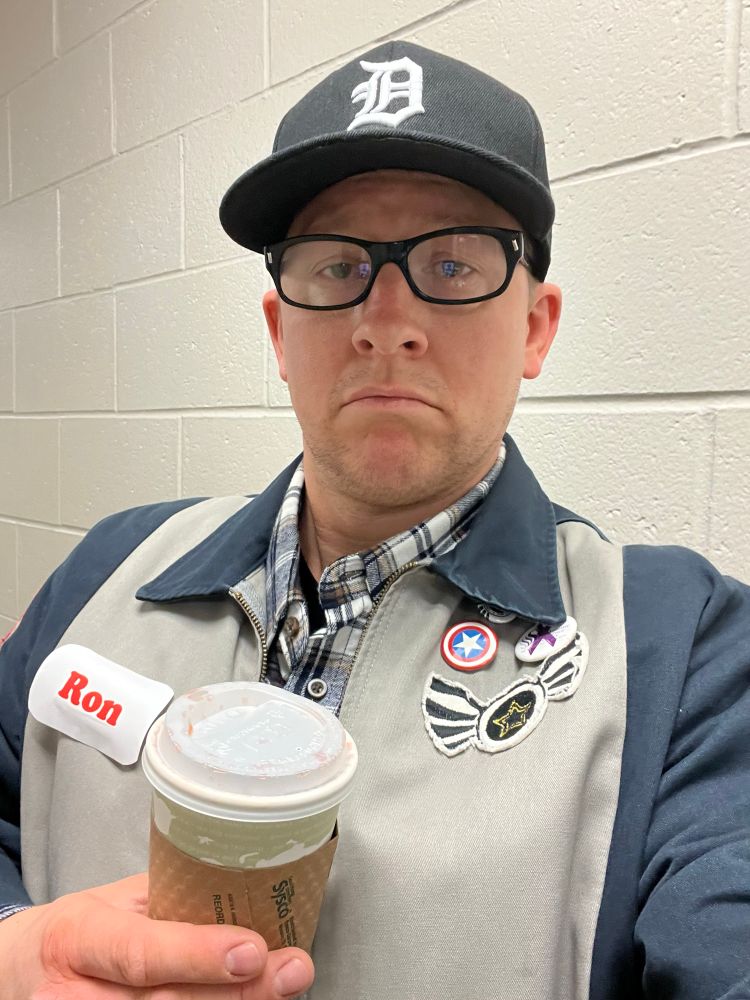 A white dude in a black Detroit ball cap, black rimmed glasses, a jacket that says Ron, holding a cup of coffee. 