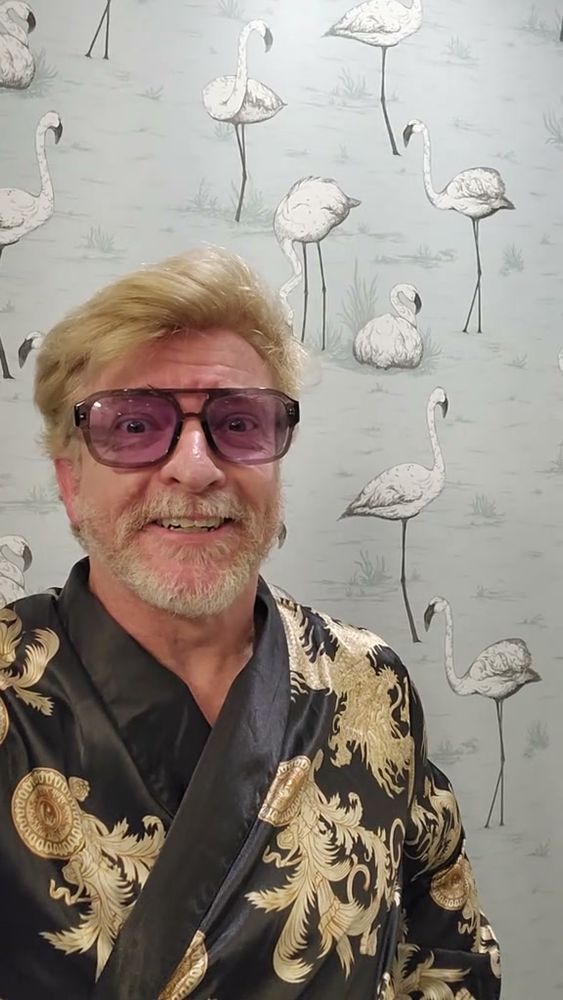 Rhys Darby looking excitedly into the camera. He's wearing a black and gold kimono and funky glasses. His beard is bearding