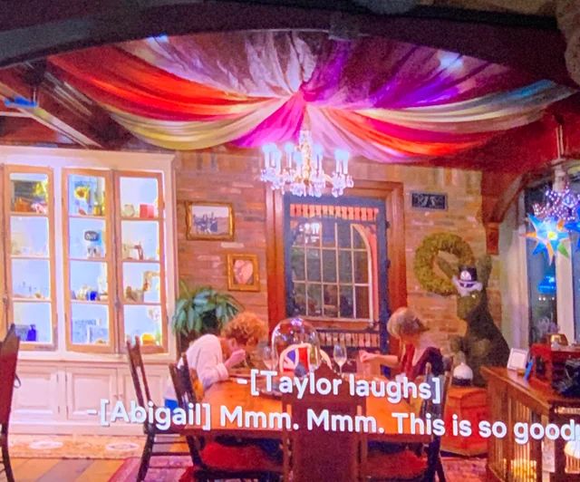 A cozy dining room is shown with colorful fabric draped across the ceiling, creating a tent-like effect. Taylor Swift and her friend Abigail are seated at the table, enjoying a meal while subtitles read “[Taylor laughs]” and “[Abigail] Mmm. Mmm. This is so good.” A decorative chandelier hangs above the table, with colorful curtains decorating the ceiling, and the room is warmly lit with personal decorations and a cabinet showcasing dishes and trinkets.