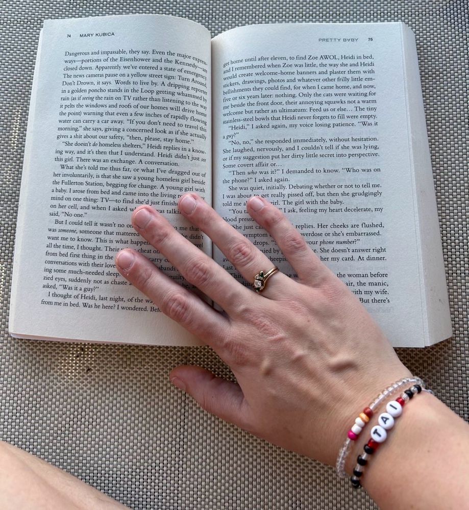 Sitting on a pool chaise, My right hand laying palm down on my open book. On my ring finger, I’m wearing a gold ring with a diamond in the middle. I’m wearing 2 friendship bracelets, one that has the lesbian flag, and the other says TAI (my dogs name) 