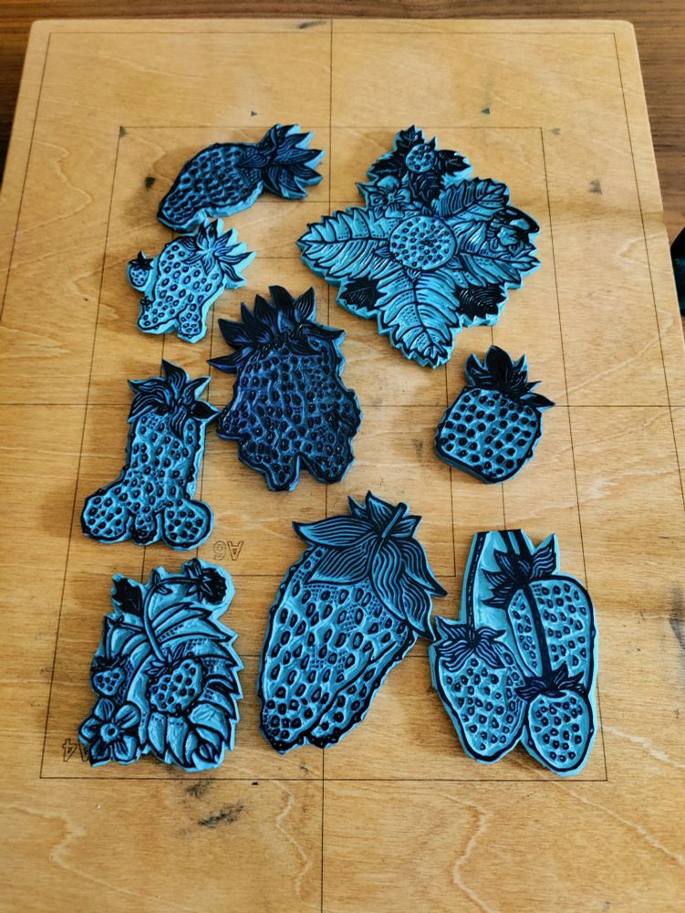 Plusieurs plaques de linogravure posées sur la presse, avec des formes et des tailles différentes. Les plaques sont bleues encrées en noir.