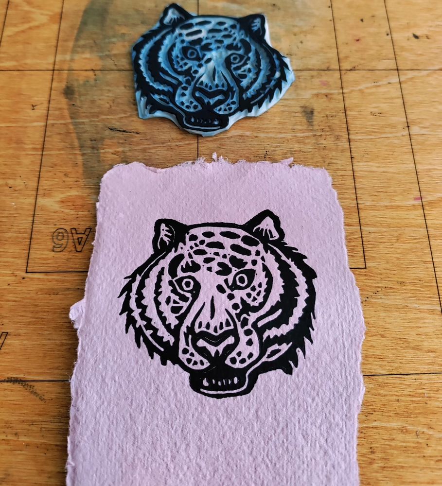 Linogravure tête de tigre avec la bouche ouverte. Imprimé sur du papier rose. On voit la plaque posée juste au dessus.