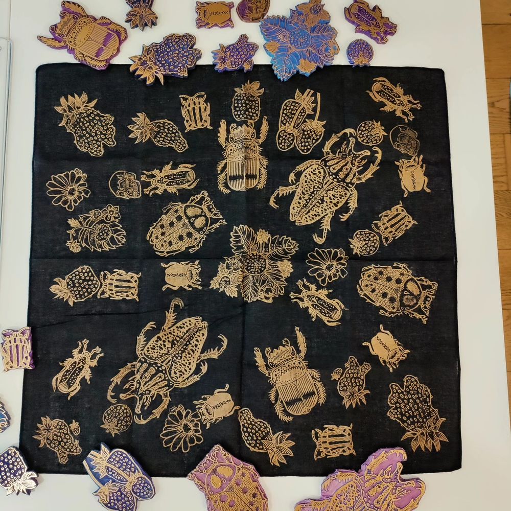 Foulard noir imprimé en or avec des motifs scarabées et fraises. Pose sur une table blanche avec les.plaques gravées 