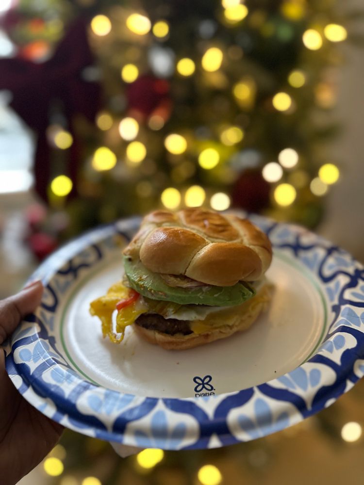 Picture of last night’s Checkers burger remixed to today’s sunrise burger: bun, patty, avocado, fried egg, on a paper plate in front of a Christmas tree. 