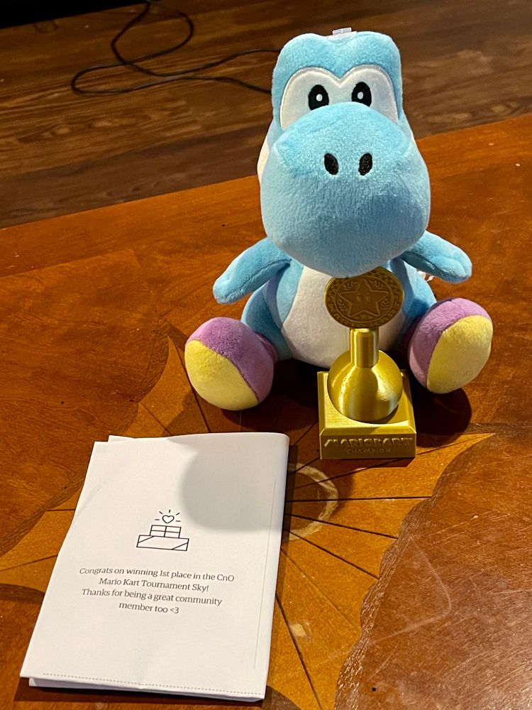 A Sky Blue Yoshi Plush “holding” a 1st Place Mario Kart trophy. There is a piece of paper next to it with a podium picture and it says “Congrats on winning 1st place in the CnO Mario Kart Tournament Sky! Thanks for being a great community member too <3”