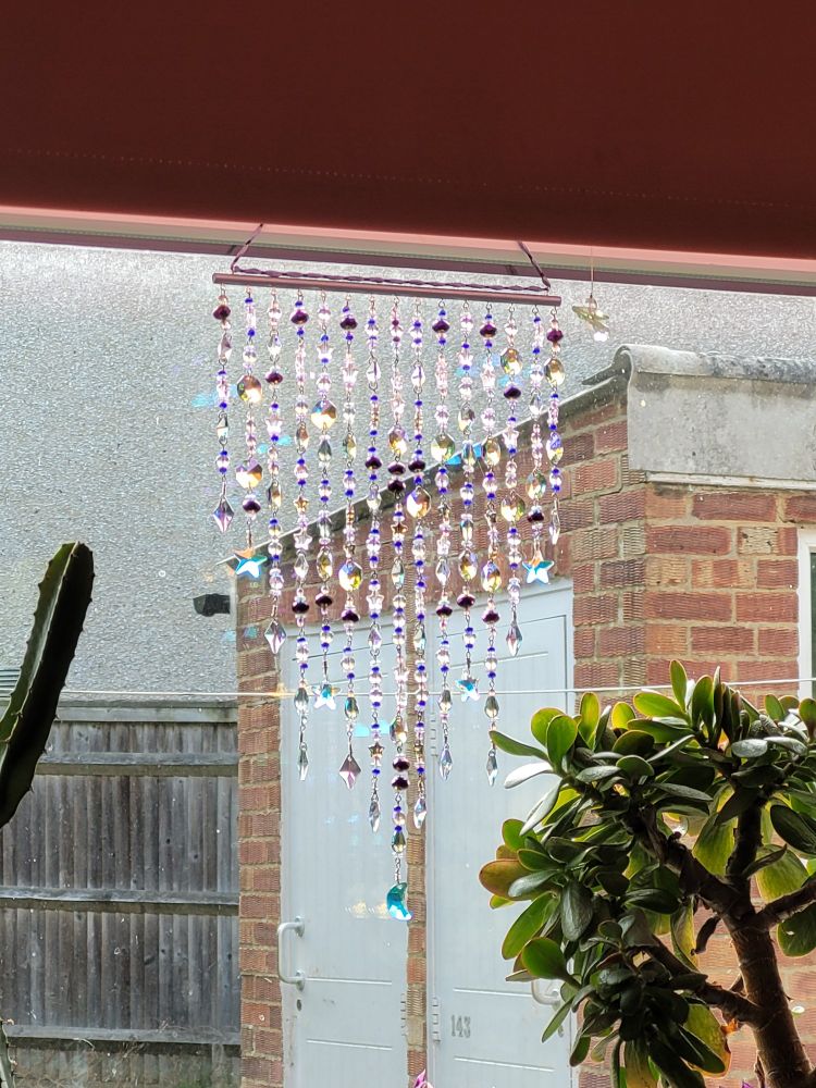 A suncatcher made of suncatching glass beads in purples, blues and whites and a few star crystals and one moon crystal. The suncatcher is pictured hanging in a window next to some plants