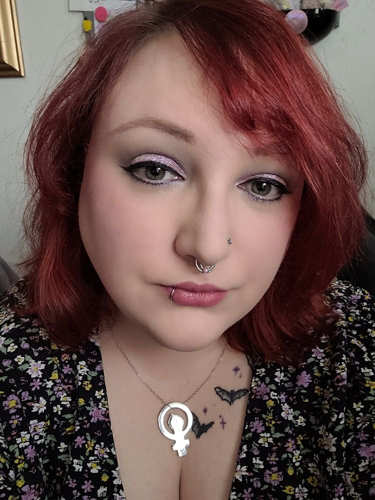 A selfie of Jess, Jess is a pale skinned woman with dyed reddish hair. She has nose and a lip piercing and wears some cute sparkly make up.
