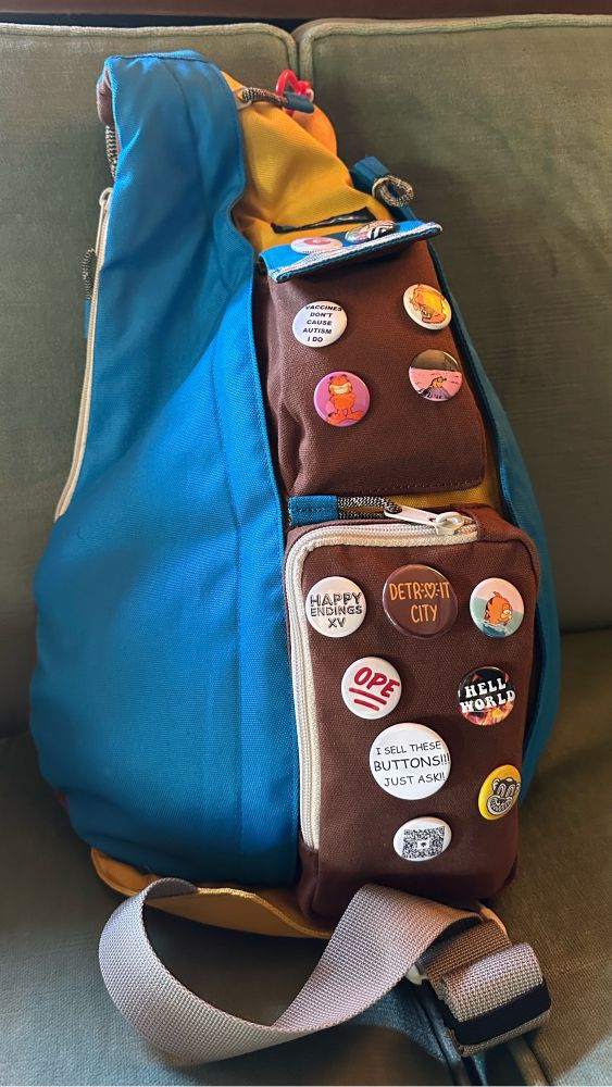 A Kavu rope bag in blue, yellow and brown color way. There are a bunch of buttons on the bag, including some Garfield ones, a hell world button, an ope button, and one that says “I SELL THESE BUTTONS!! Just ask!”