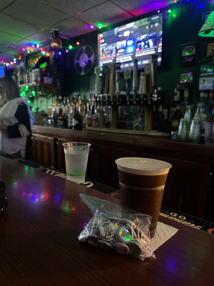 A picture of a small bag of buttons including front of a Guinness, which is sitting on a bar top. The back of the bar is a ton of liquor as is custom for a bar