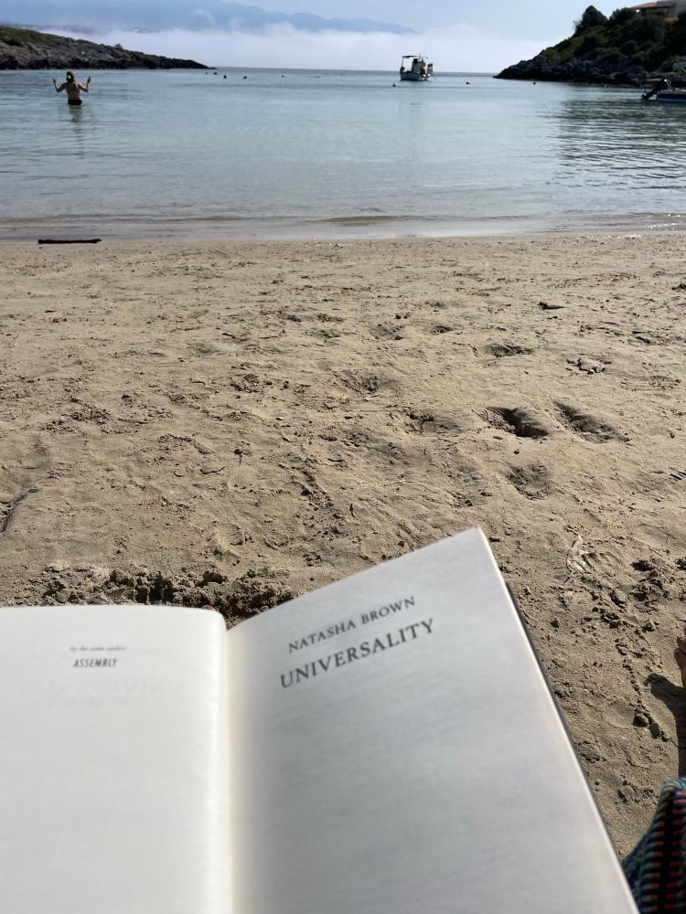 Reading Universality by Natasha Brown on a sandy beach, with a calm blue sea in the background. 