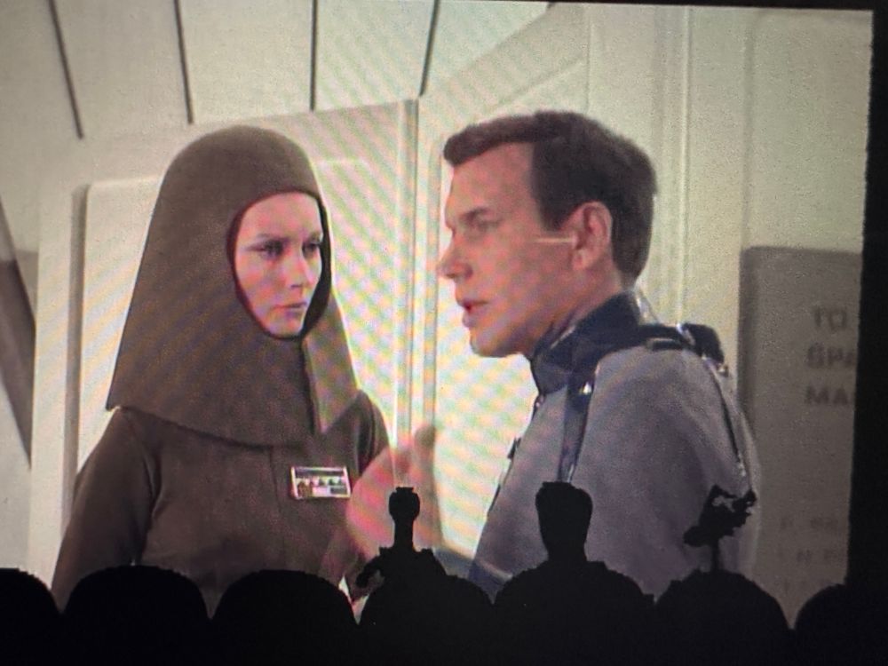 A still from Moon Zero two featuring a woman with a foam piece of headgear that is round on top and flares at the bottom to her shoulders, making her appear to be wearing an oversized helmet. The hat and suit are brown, with two rows of buttons on the chest. She’s talking to a man in a grey suit. 