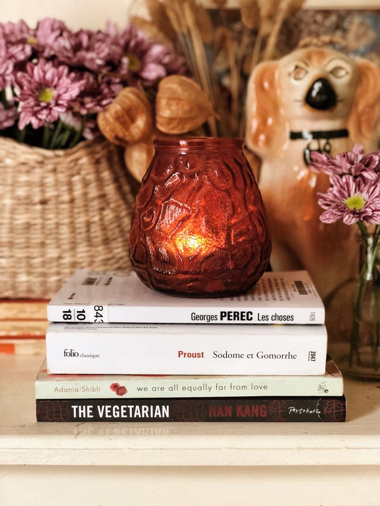 A stack of books, purple mums in a basket, a porcelain dog and an orange candle holder on a mantelpiece. The books in the stack are: Les Choses by Georges Perec, Sodome et Gomorrhe by Marcel Proust, We Are All Equally Far From Love by Adania Shibli and The Vegetarian by Han Kang.