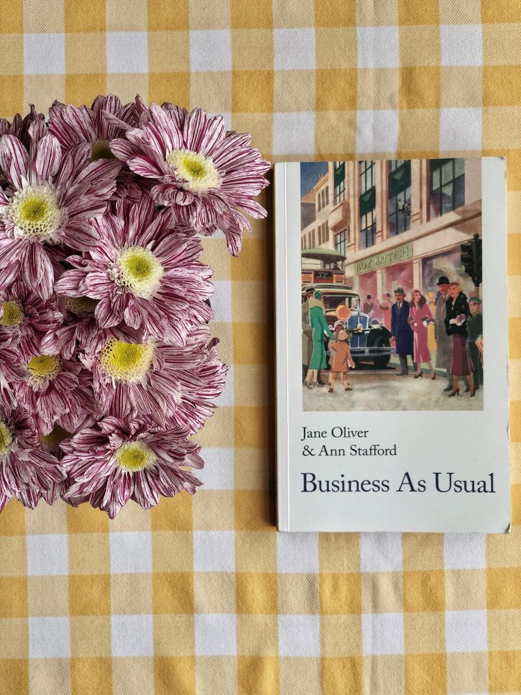 On a yellow gingham tablecloth, a bunch of purple chrysanthemums and a book: Business as Usual by Jane Oliver & Ann Stafford