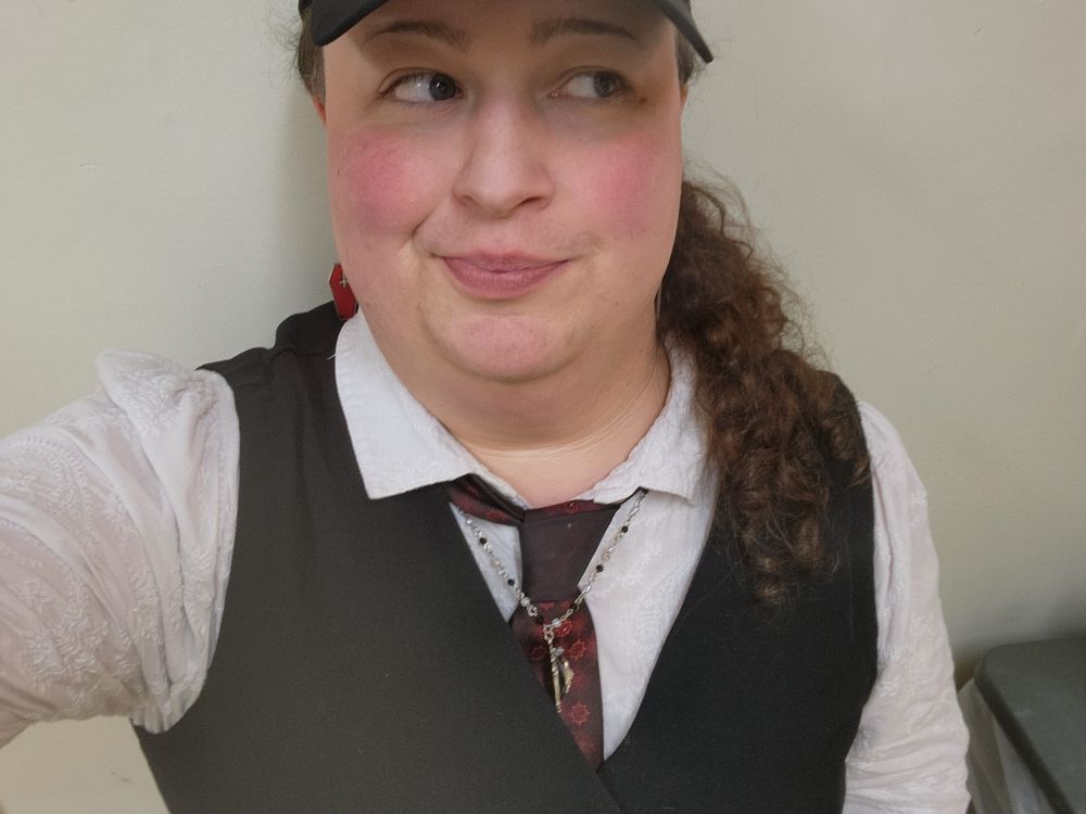 A woman in a black cap, white collared button up blouse, red vintage tie, black vest with long curly hair looking coquettishly off to the right of the image while smirking.