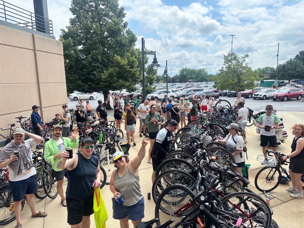 A bunch of people at the bike racks waving at the camera, some holding drinks and some holding popsicles