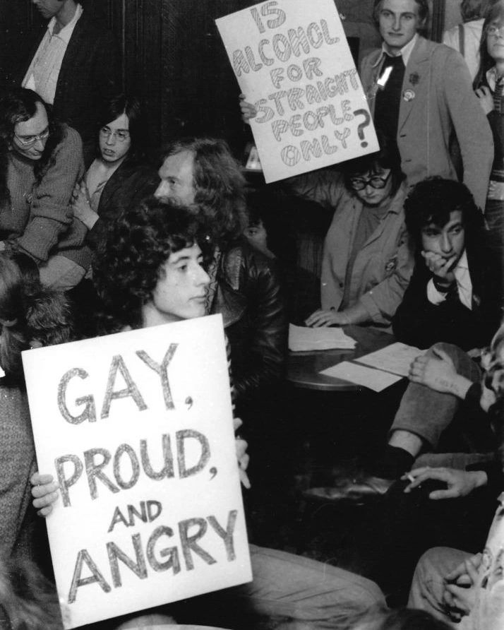 Candid photo of a protest/demonstration from the 70's; a person in the foreground is holding a sign that says, "Gay, proud, and angry."