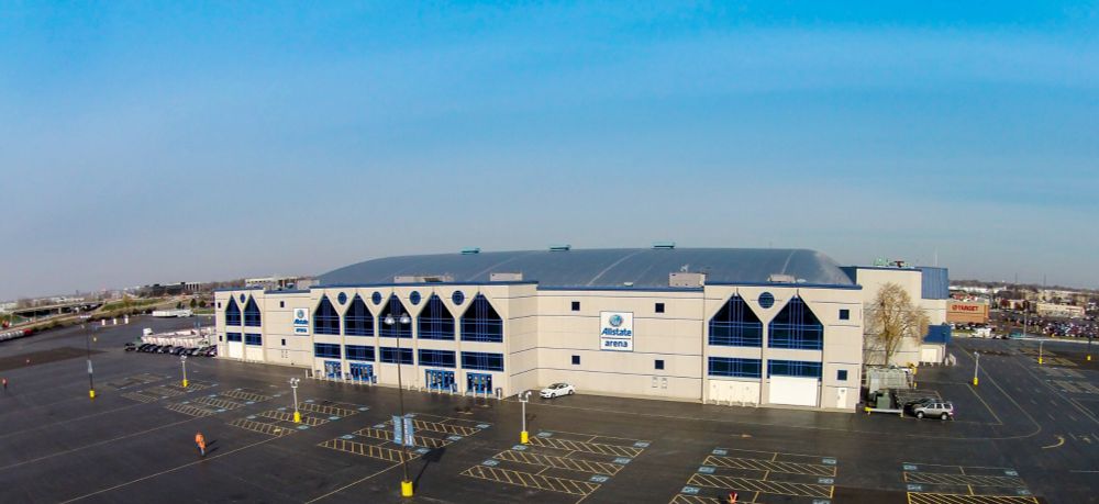 An exterior picture of the Allstate Arena, a large building used for sports and concerts in Rosemont, Illinois. It's a large tan building with a blue roof, and large trapezoidal black windows. There are also a few doors and a lot of parking lot showing in the picture. If you squint at the left, you can see the highway with the one goddamn exit that you can take to get to this arena, which creates a nightmarish hellscape in the parking lot. If you attend an event here, make sure you pee before you try to leave.