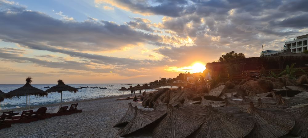 une plage en Albanie au coucher du soleil, avec un TAS de parasols en genre de truc végétal empilés par terre