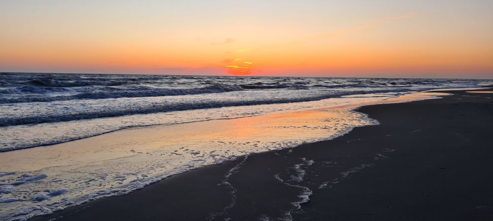 la mer qui déferle sur une plage de sable au soleil couchant, la pellicule d'eau au bord reflète le ciel orange et on voit une petie vague arriver qui fait toute la longueur de la plage