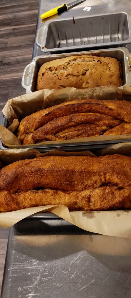 An image of four loaf pans on a silver table. There's a yellow and black handled knife at the top of the image. The top load pan is empty. It had a date nut bread in it l, but I pulled it out to cut it. 

The next one down had another date nut bread in it. It's golden brown with a cracked top dome

Below that one is two more loaf pans, they are longer than. The top two, and both contain a loaf of cinnamon bread. The upper cinnamon bread was over scored and cut through some of the layers. When baked it puffed into a vague "labia" shape. 

The one below that one looks normal. Mostly.