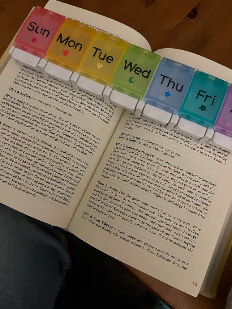 A book about flavor combinations, opened to the “Olive” chapter (Olive & Anise; Olive & Bacon, etc), is held open by a rainbow-multicolored 7-day pill organizer 