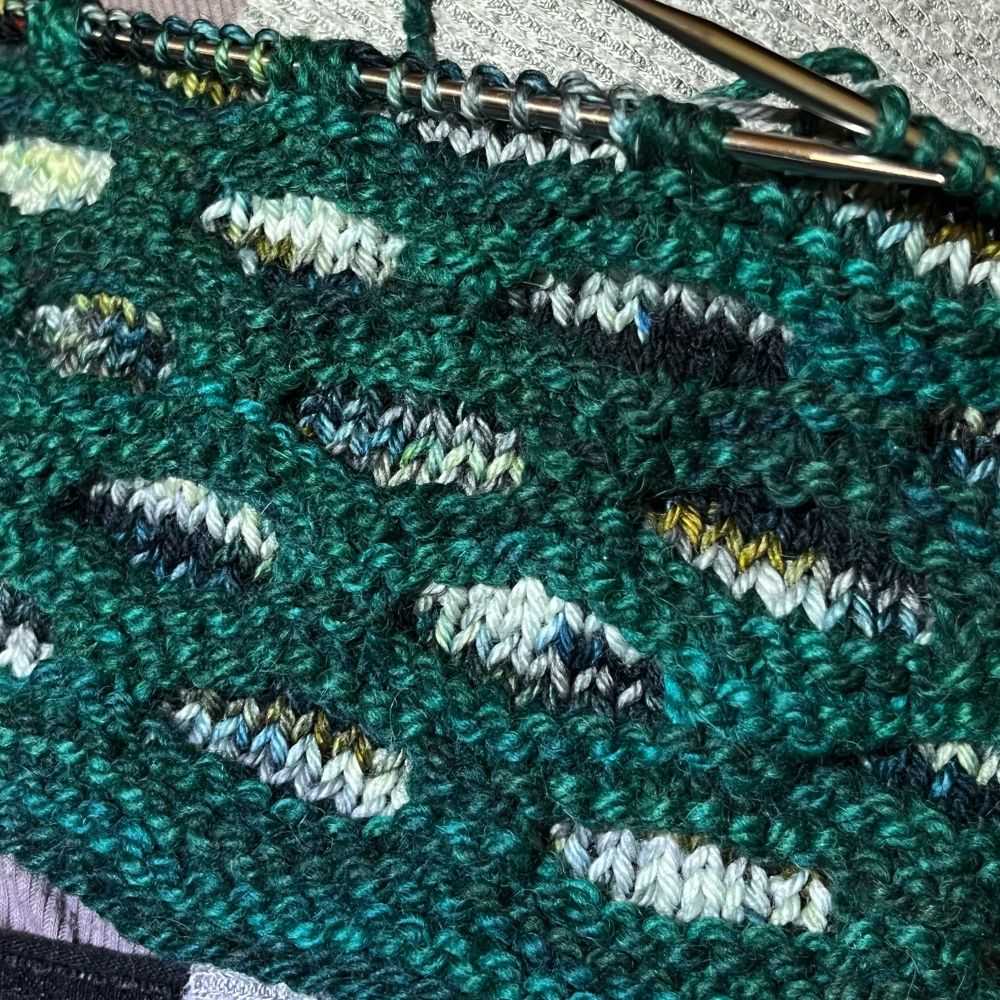 Close up photo of a knitted cowl in progress, with lozenge-shaped openings of white, black, blues, greys and greens appearing in random patterns, and a foreground of bluish forest green.