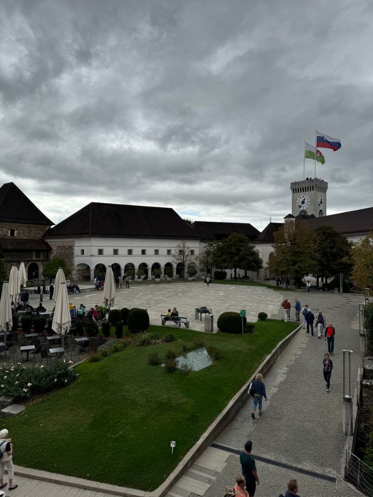 Blick in den Innenhof der Burg über Ljubljana. Sie ist sehr schön restauriert. Im Vordergrund ist eine gepflegte Grünfläche die von einem breiten Weg umschlossen ist. Im Hintergrund sieht man einen Teil der Burg und einen Burgturm auf dessen Dach die Fahne von Ljubljana und die Landesfahne von Slowenien im Wind flattern