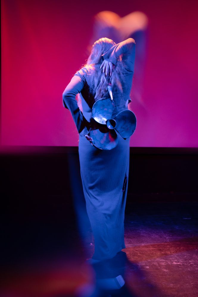 Blonde woman in costume performing on stage in SNOLIGOSTER. Theatrical lighting and surreal in-camera effects create a dreamlike, satirical atmosphere. All images captured live using a Reflect Lens filter.