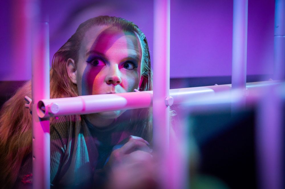Blonde woman in costume performing on stage in SNOLIGOSTER. Theatrical lighting and surreal in-camera effects create a dreamlike, satirical atmosphere. All images captured live using a Reflect Lens filter.