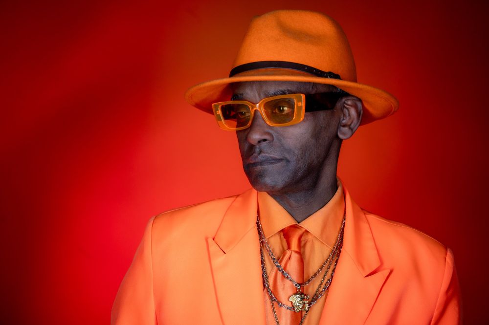 Portrait of a stylish man wearing a bright orange suit, matching fedora, and amber-tinted sunglasses. He wears layered silver chains over an orange shirt and tie. The background is a smooth gradient of warm red and orange tones, enhancing the vibrant, monochrome colour palette.