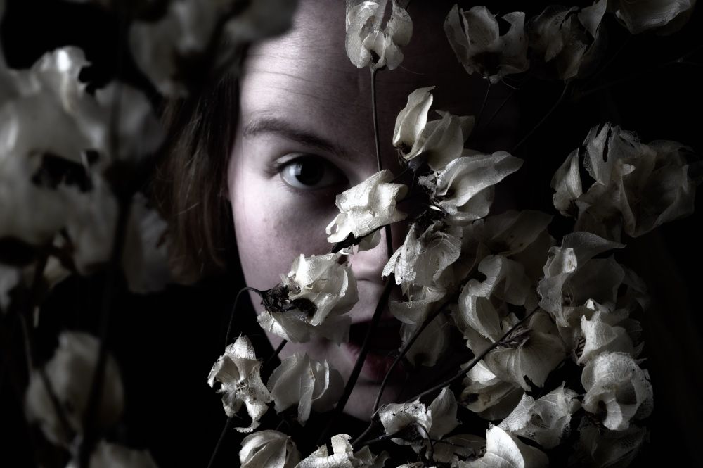  A close-up portrait of a woman with blond hair, her face partially hidden behind a cluster of white dried flowers. One large eye stares directly into the camera, framed by petals and shadow. The lighting is low-key and moody, with deep shadows and a cool, muted tone.