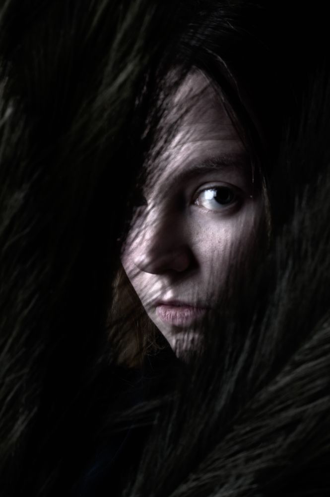 A moody close-up of a woman’s face peeking through what appears to be dark, soft grass or feathery foliage. Her eye is lit dramatically, emerging from the shadows, while the rest of the frame is enveloped in rich darkness. The image feels mysterious and intimate.